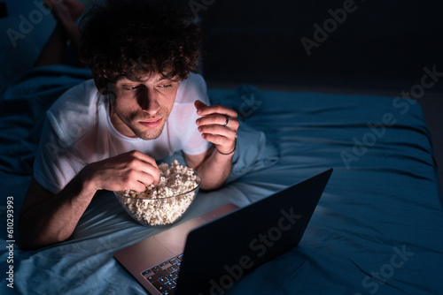 Canvastavla Young man eating popcorn and watching movie on laptop computer at night at home