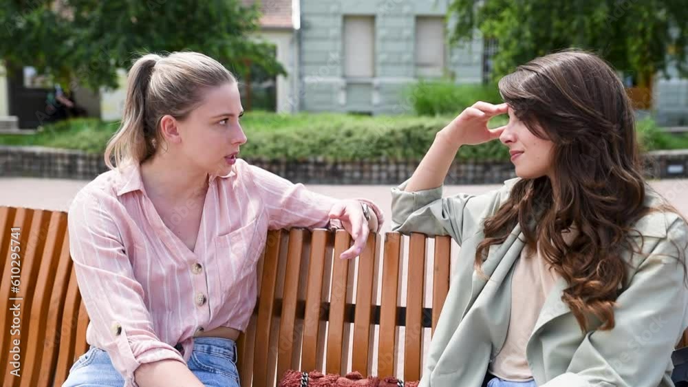 Two best friends talking outside, sitting on a bench in a park. Women friendship on sunny day in the city conversation and happiness. Business colleagues at break talking gossiping.