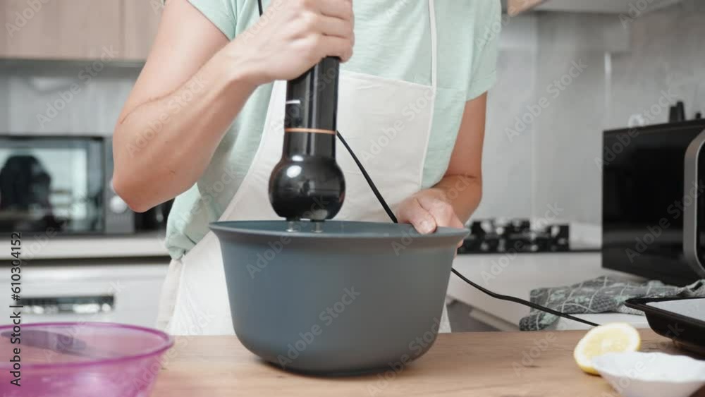 In a slowed-down shot, a woman is using a black hand mixer to whisk the ingredients in a bowl. The camera moves in a circular motion around her. In a slowed-down shot, a woman is using a black hand mixer to whisk the ingredients in a bowl. The camera moves in a circular motion around her.
