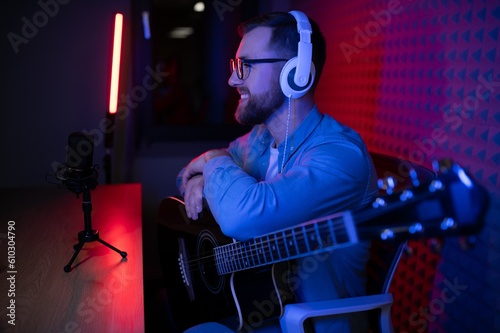 Young Caucasian male singer in headphones hold play guitar record new single on smartphone at home studio. Millennial man artist use musical instrument sing shoot music video on cellphone camera.