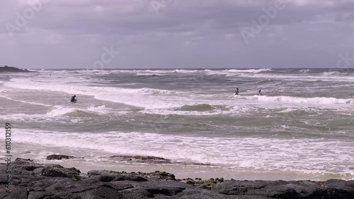 People play in rough surf by sandy beach on overcast day. Waves are wild, headland in background, rocks in foreground