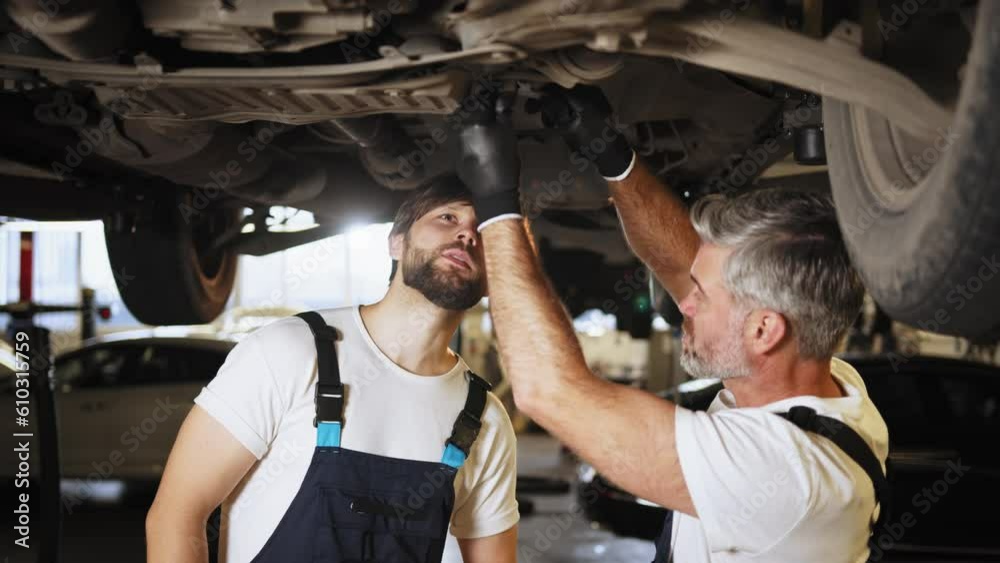 Two auto mechanics men in uniform at car suspension repair work. Vehicle raised on lift at