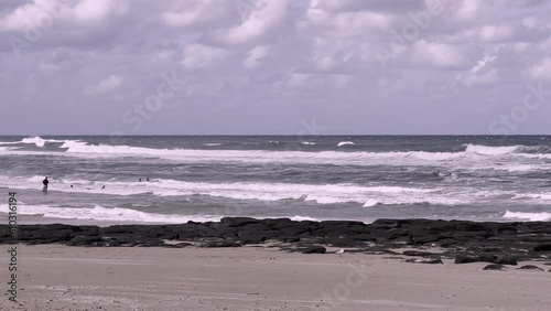 People enter wild surf at beach on overcast day. Shore is both sandy and rocky