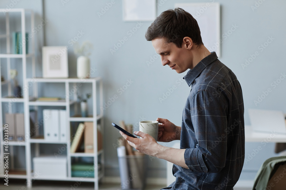 Side view portrait of smiling young man using smartphone in office at coffee break, copy space
