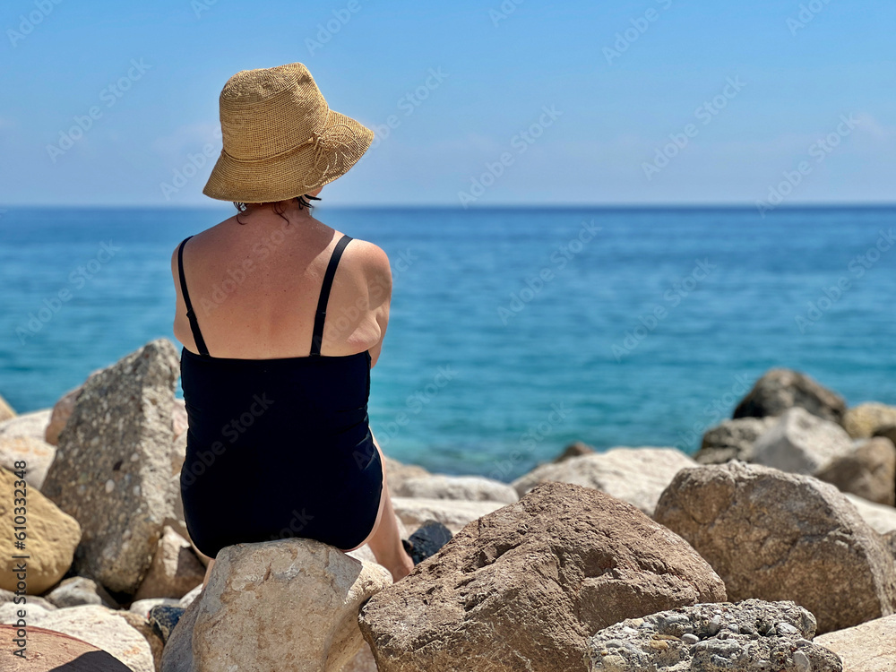 Senior woman in black one piece bathing suit sitting on the rocks with her back to the viewer, gazing at the sea in front of her