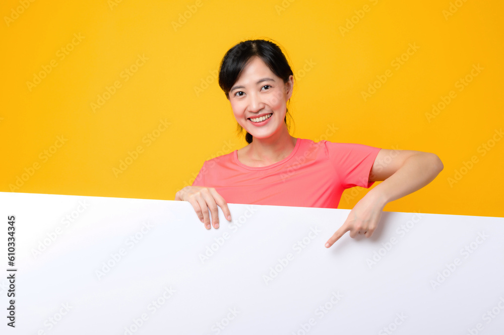 young asian sports fitness woman happy smile wearing pink sportswear standing behind the white blank banner or empty space advertisement board against yellow background. wellbeing lifestyle concept. young asian sports fitness woman happy smile wearing pink sportswear standing behind the white blank banner or empty space advertisement board against yellow background. wellbeing lifestyle concept.