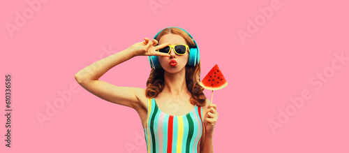 Summer portrait of stylish woman in headphones listening to music blowing her lips with juicy lollipop or ice cream shaped slice watermelon, female model posing on pink background