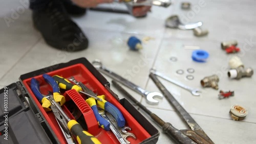 A male master plumber repairs a disassembled faucet from a bathroom, changes the cartridge and gasket. On the ceramic floor is a toolbox and a dismantled faucet.