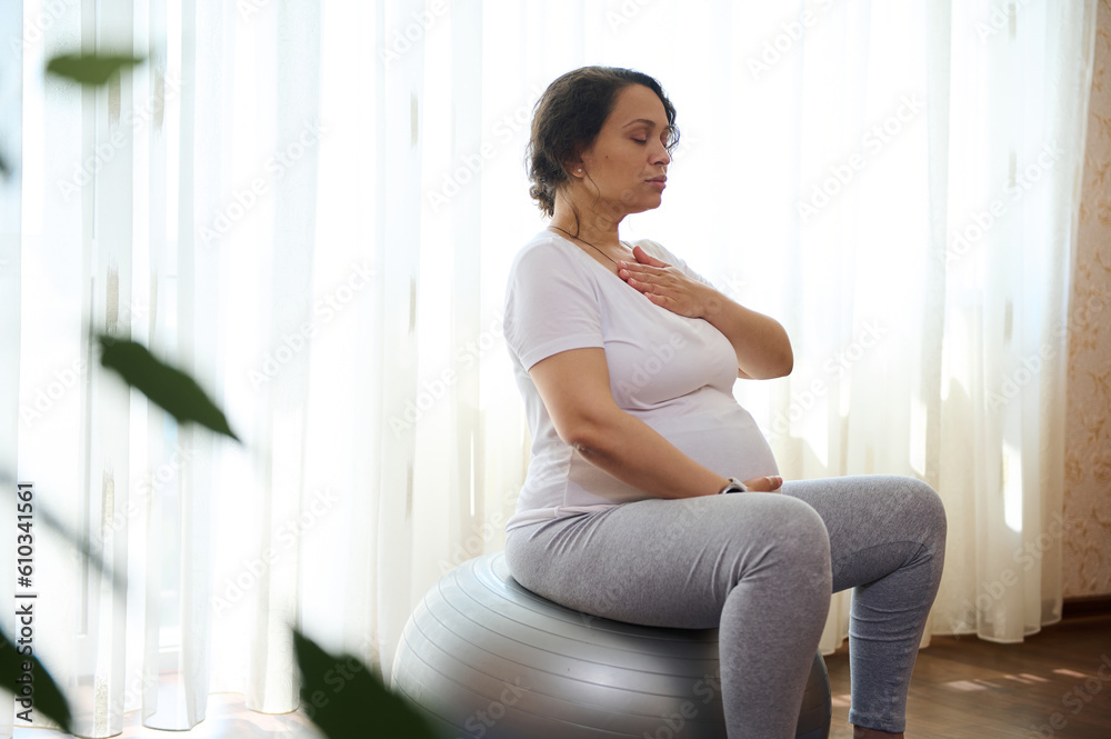 Pregnant adult woman in sportswear, sitting on a fit ball, doing meditation and pranayama breathing exercises for healthy pregnancy, preparing body for childbirth, putting hands of chest and belly Pregnant adult woman in sportswear, sitting on a fit ball, doing meditation and pranayama breathing exercises for healthy pregnancy, preparing body for childbirth, putting hands of chest and belly