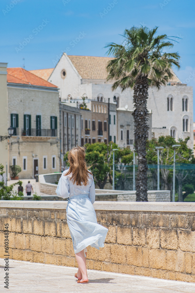 Fototapeta premium Blond hair girl walking on the ancient walls with Pontifical Basilica or Church of Saint Nicholas and palm trees on background in Bari old town, Puglia or Apulia, vertical