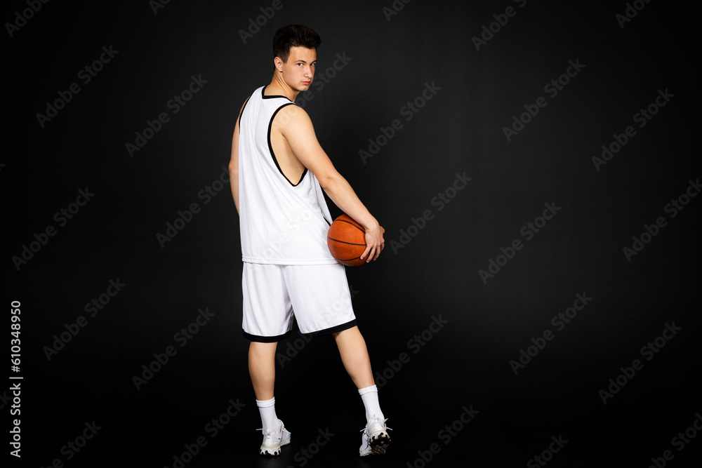 Handsome young basketball player in white sportswear holds a ball. Isolated on black background. view from behind