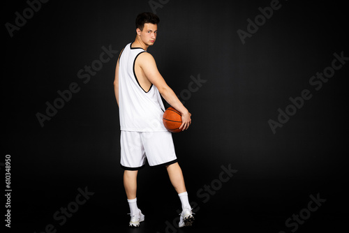 Handsome young basketball player in white sportswear holds a ball. Isolated on black background. view from behind © Georgi