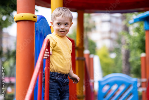 Sweet blond kid with Down syndrome standing at climber smiling and holding on to railing positive little boy in yellow t-shirt enjoys spending time at usual playground