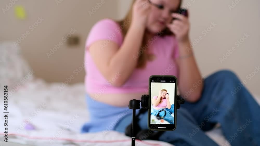 TikTok beauty blogger recording process of makeup on camera to show followers teenage girl painting lashes with mascara sitting on bed behind phone on tripod TikTok beauty blogger recording process of makeup on camera to show followers teenage girl painting lashes with mascara sitting on bed behind phone on tripod
