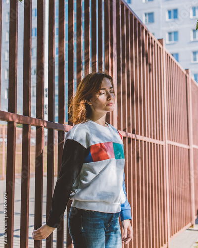 The girl is standing with her back to the fence with her eyes closed
