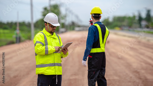 Asian engineer holding tablet and foreman using walkie talkie to control road construction, against backdrop of road under construction