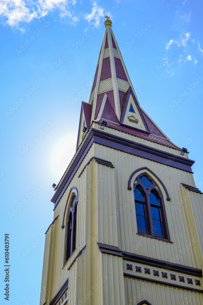 A yellow colored tall wooden Catholic square shaped steeple of a religious building. The exterior decorative clerestory windows have red trim. The peaked wood roof has decorative molding designs.  