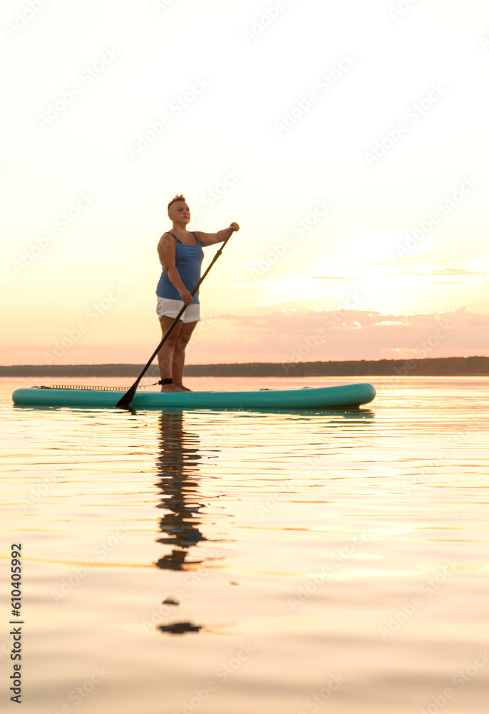 A woman in mohawk shorts stands on a SUP board with a paddle at sunset in a lake against the backdrop of a sunset sky and water.