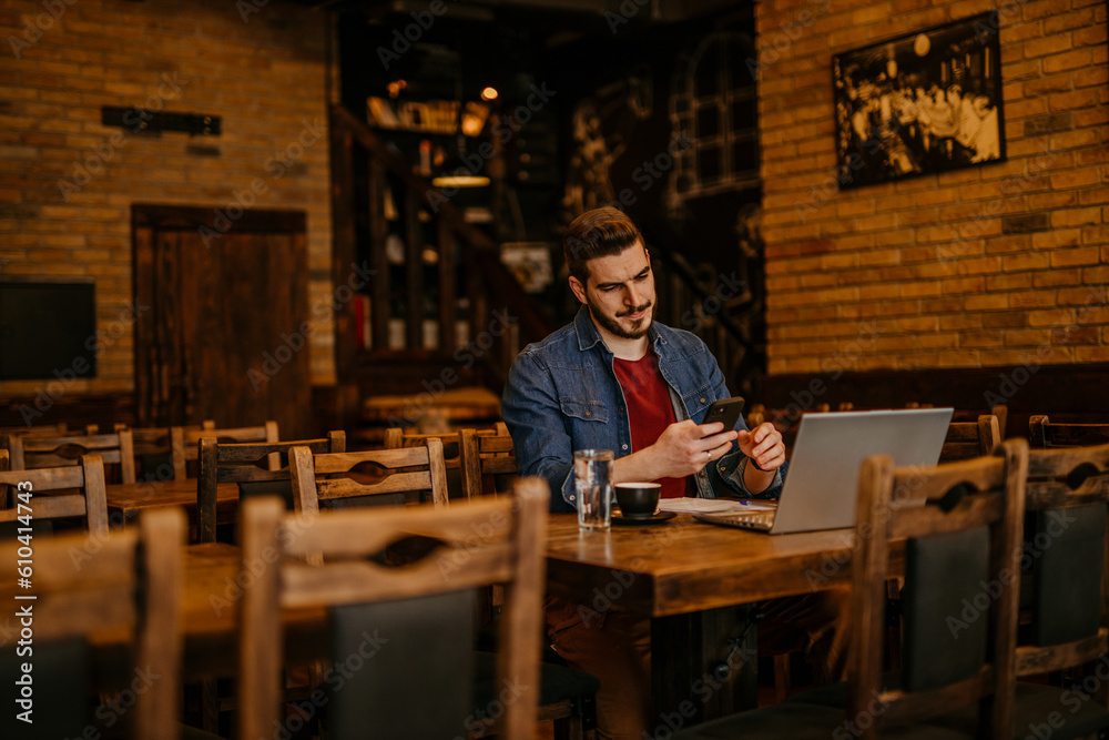Young businessman owner or manager of a cozy bar and cafeteria, using a phone and a laptop computer to prepare everything for the upcoming day