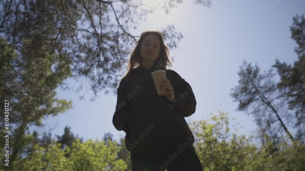 Portrait of attractive cheerful girl drinking hot latte drink, relaxing, walking outdoors outdoors in the park. A girl in a brown sweatshirt walks in a city park and drinks coffee. Sunny weather. Portrait of attractive cheerful girl drinking hot latte drink, relaxing, walking outdoors outdoors in the park. A girl in a brown sweatshirt walks in a city park and drinks coffee. Sunny weather.