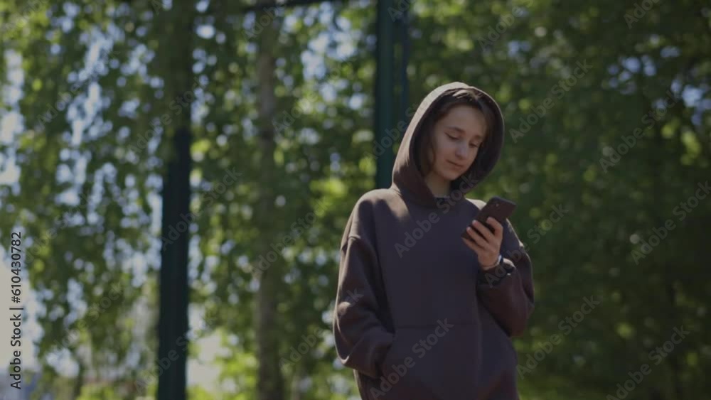Attractive girl holding a mobile phone in her hands on the street, on a sunny summer day, side view. The girl looks at the smartphone, on the street sports ground. Social networks, online, chatting. Attractive girl holding a mobile phone in her hands on the street, on a sunny summer day, side view. The girl looks at the smartphone, on the street sports ground. Social networks, online, chatting.