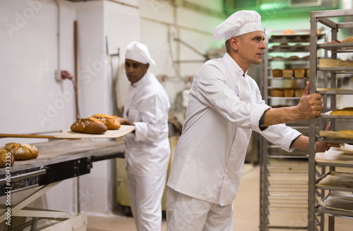 man in chefs uniform rolling trolley with bread in bakery