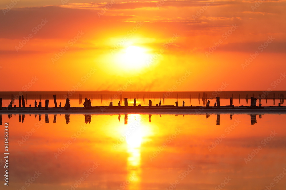 Orange sunset, dramatic and majestic sky over beach with sun reflection in water. Wood posts for salt extraction of extremely salty lake, Ukraine. Summer season, idyllic peaceful dusk sunlight