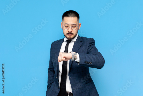 successful asian businessman in suit and glasses is late and looks at his watch on blue isolated background photo