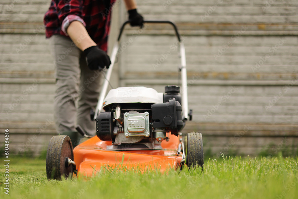 Obraz premium Unrecognisable gardener man in protective gloves starts the lawnmower before cutting green grass lawn in his backyard. Man with motorised lawnmower cares for landscaping lawn