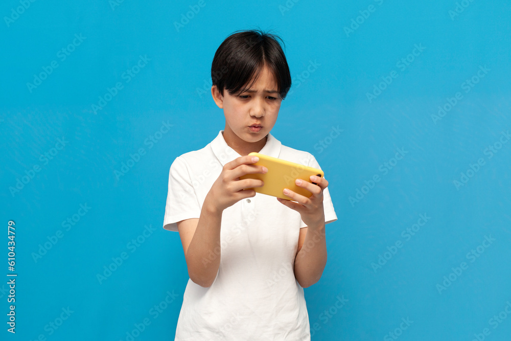asian boy of twelve years playing video game on smartphone on blue isolated background asian boy of twelve years playing video game on smartphone on blue isolated background