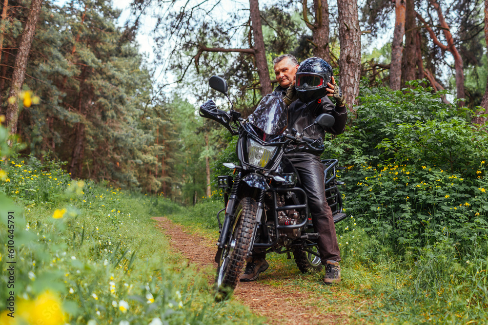 Senior biker taking helmet off after riding motobike in summer forest. Retired man wearing leather clothes. Hobby