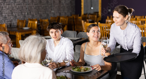 Elderly couple and young married spouses eating in restaurant. Cafe, festive lunch is event for narrow circle of close friends. Friendly waitress brought glass of wine for young girl visitor