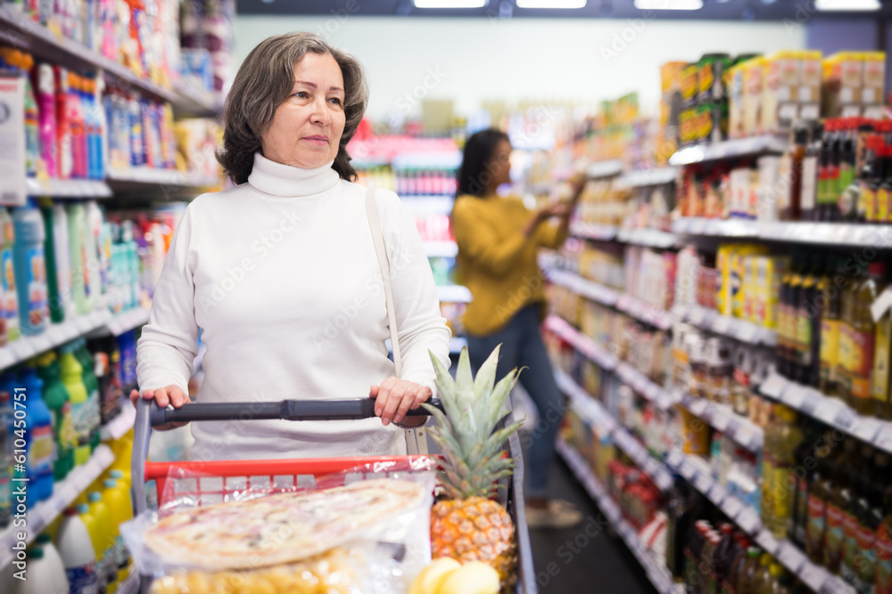 Pensive older woman shopping at store, walking with cart among shelves and choosing products