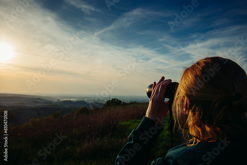 A hiker girl looks through binoculars at nature and birds standing on the top of a mountain in the bright rays of setting sun against the background of beautiful spring nature and cloudy blue sky