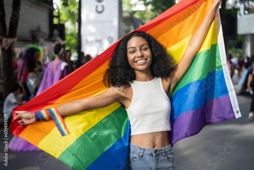 Young beautiful woman posing with rainbow flag at city street. She looking to camera with attractive smiling.