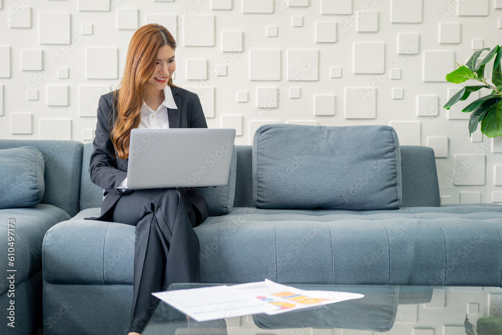 Image of a young beautiful Asian Business woman smiling while working with a laptop and talking to work doing math finance on an office desk, tax, report, accounting, statistics, and research concepts Image of a young beautiful Asian Business woman smiling while working with a laptop and talking to work doing math finance on an office desk, tax, report, accounting, statistics, and research concepts