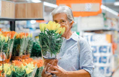 Senior woman wearing glasses shopping in the garden department of the supermarket. Mature woman picks sniffs yellow flowers. Consumerism concept photo