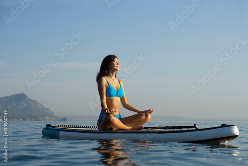 Asian beautiful girl in a blue bikini doing ugha sits in a lotus position with her eyes closed and meditates on a water sports board in the ocean against the background of a sunset. Woman on Sup.