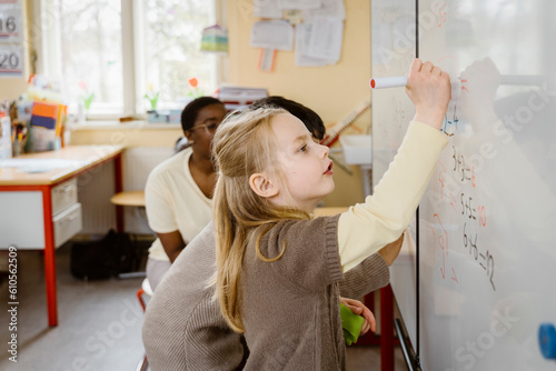 Blond girl solving maths problem while writing on whiteboard in classroom photo