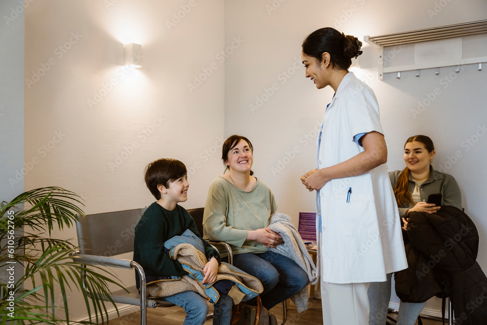 Side view of female healthcare worker wearing lab coat talking to boy sitting on chair in waiting room at hospital Side view of female healthcare worker wearing lab coat talking to boy sitting on chair in waiting room at hospital