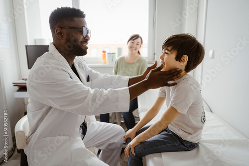 Smiling male pediatrician examining neck of boy sitting on bed at healthcare center photo