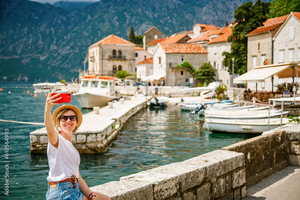 Tourist woman making a selfie sitting on the embankment of the small town Perast