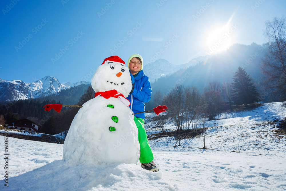Obraz premium Young boy in winter outfit stand with freshly build snowman