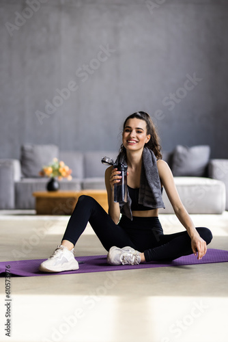 Young woman in sportswear drinking water while sitting after exercising on a yoga mat at home.