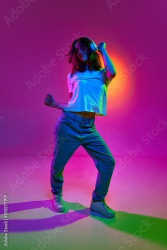 Portrait of a young girl wearing jeans, white t-shirt and headphones listening to music, dancing with pleasure over pink background in neon light