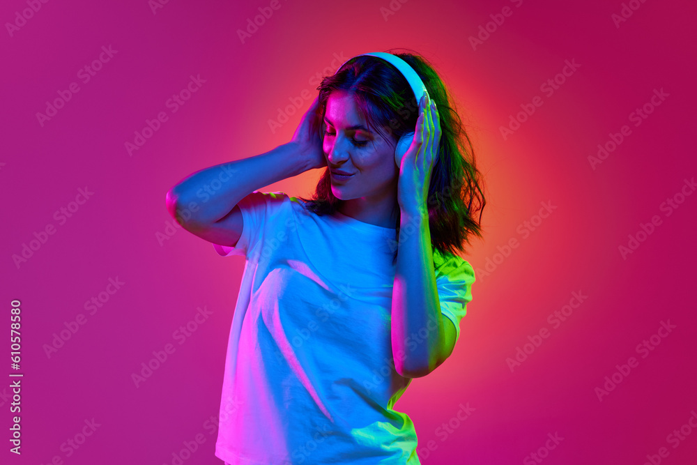Image with young, positive brunette girl, DJ wearing headphones creating music beats and smiling over pink studio background in neon light Image with young, positive brunette girl, DJ wearing headphones creating music beats and smiling over pink studio background in neon light
