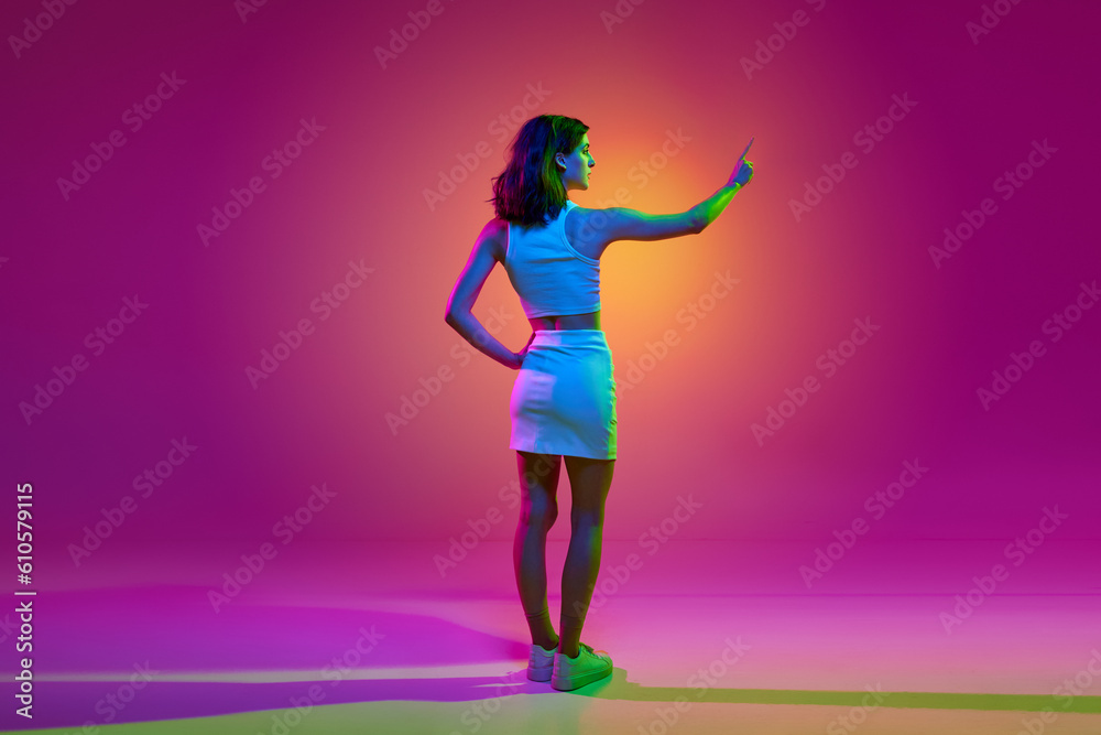 Portrait of a young girl standing and touching an imaginary screen over pink color studio background in neon light. Concept of gadgets, online education