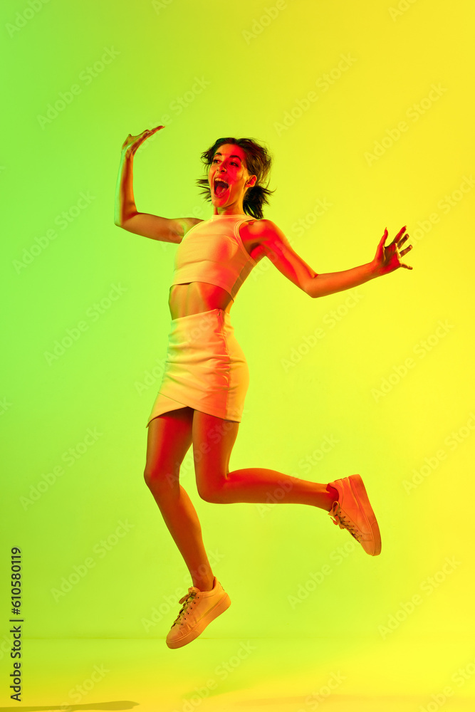 Funny portrait of emotive, young girl wearing skirt and top jumping up over acid green color studio background in neon light. Freedom