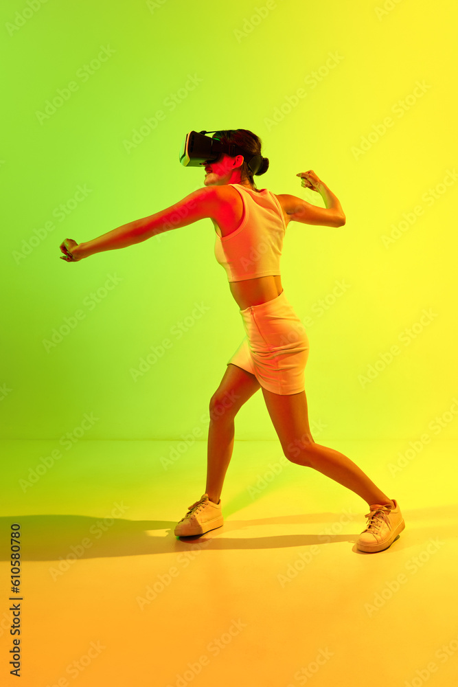 Girl wearing VR headset glasses playing game over acid green and yellow background. Youth, virtual lifestyle of future. Virtual reality fight