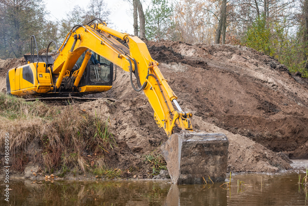 Excavator digging artificial water basin. Long arm of an excavator spreading banks of a pond, making it bigger Excavator digging artificial water basin. Long arm of an excavator spreading banks of a pond, making it bigger